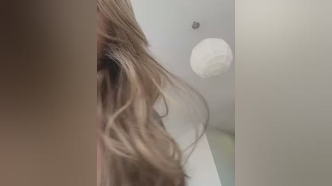 Media: Video of a woman's head with long, wavy blonde hair partially covering her face, seen from the side. Background shows a white ceiling with a round light fixture.