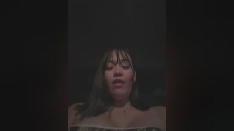 Media: Video of a young woman with medium-length, wet hair, wearing a white off-shoulder top, with a dark, blurred background. She appears to be in a state of distress, with her eyes closed and mouth slightly open.