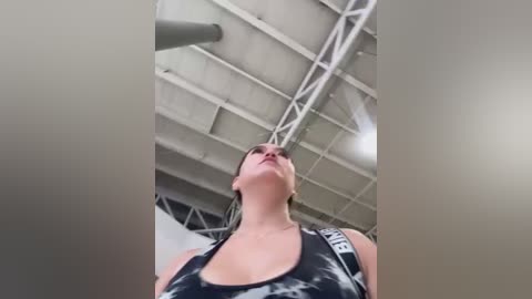 Media: Video of a woman with light skin and blonde hair, wearing a black and white sports bra, looking up at a gym ceiling with exposed beams and lights.