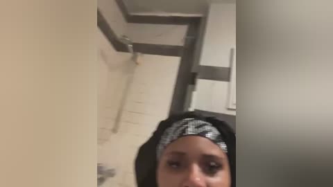 Media: Video of a woman with medium skin tone, wearing a black and white patterned hijab, standing in a beige-tiled bathroom with a sink and mirror, holding a smartphone.