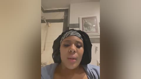 Media: A video of a Black woman with medium brown skin, wearing a black headscarf and a light blue shirt, standing in a bathroom with white walls and a framed painting.