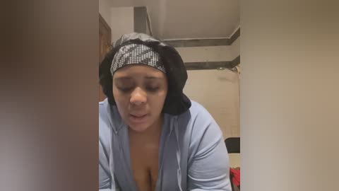 Media: A video shows a woman with a light brown complexion, wearing a black headscarf and a light blue hoodie, her eyes closed, looking tired, standing in a dimly lit room with beige walls and a ceiling fan.