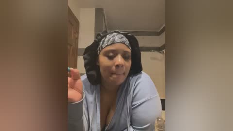 Media: Video of a light-skinned Black woman with a large bust, wearing a light blue shirt, black headscarf, and black headphones. She's holding a phone, smiling, in a bathroom with beige walls and a wooden door.