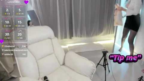 Media: Video of a woman in a white dress and black shorts, standing in a white room with a white couch and a camera on a tripod.