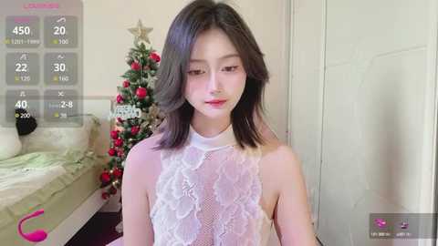 Media: A video of a young East Asian woman with shoulder-length black hair, wearing a sheer white lace top, standing in a bedroom with a decorated Christmas tree.