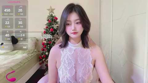 Media: Video of a young Asian woman with shoulder-length black hair, wearing a sheer white lace top, standing in a cozy room with a decorated Christmas tree and a bed in the background.