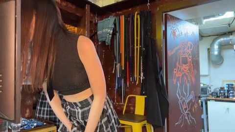 Media: Video of a young woman with long, straight brown hair, wearing a black crop top and a plaid skirt, standing in a cluttered, dimly lit workshop with various tools and a motorcycle helmet hanging on the wall.