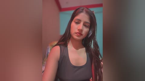 Media: Video of a young South Asian woman with long, dark hair, wearing a black tank top, standing indoors with a blurred background.