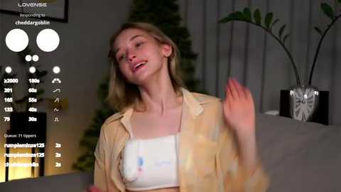 Media: Video of a young woman with fair skin, light blonde hair, and a white top, smiling while waving, in a dimly lit room with a potted plant.