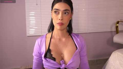 Media: Video of a young woman with olive skin, dark hair, and medium-sized breasts, wearing a purple blouse and black lace bra, standing in a pastel-painted room with white blinds.