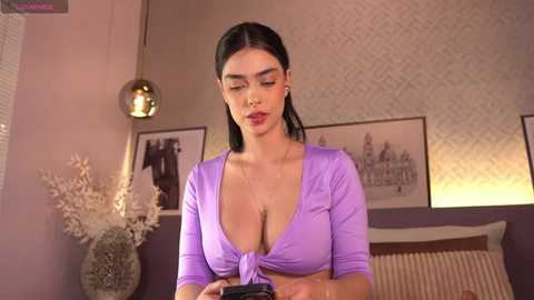 Media: Video of a brunette woman in a purple crop top, sitting on a bed, holding a camera, with modern decor in the background.