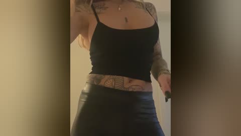 Media: Video of a tattooed woman in a black crop top and tight black leather pants, standing indoors with dim lighting.