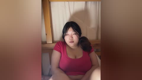 Media: A video of a plus-sized East Asian woman with long black hair, wearing a pink T-shirt, sitting on a bed with a wooden frame. She has a relaxed, slightly open-mouthed expression. The background is plain with beige walls and curtains.