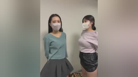 Media: Video of two East Asian women wearing white masks, teal sweaters, and high-waisted skirts. One is in a teal skirt, the other in a black skirt. They stand in a plain, light-colored room, with soft lighting.