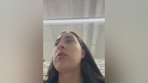 Media: Video of a young woman with long, dark hair, looking up thoughtfully, in an industrial setting with white walls, exposed pipes, and fluorescent lighting.