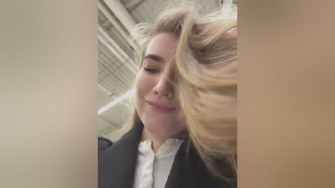 Media: A video of a blonde woman with fair skin and a septum piercing, wearing a black coat over a white blouse, laughing and tilting her head back in a bright, industrial setting.