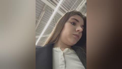 Media: Video of a young woman with light skin and straight, shoulder-length brown hair, wearing a white blouse and dark jacket, looking contemplative under a high, industrial ceiling with exposed beams.