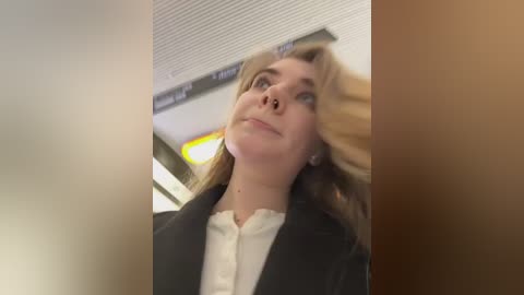 Media: Video of a Caucasian woman with blonde hair, wearing a black jacket over a white blouse, looking upward, in an indoor setting with ceiling lights and blurred background.