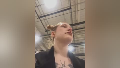 Media: Video of a young woman with pale skin and blonde hair styled in a high bun, wearing a black blazer and a revealing top showing a tattoo of deer antlers. She looks upward, captured from a low angle.