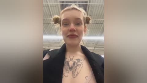 Media: Video of a fair-skinned woman with blonde hair in two buns, wearing a black jacket. She has a detailed butterfly tattoo on her chest. The background features a metal ceiling.