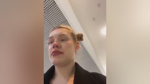 Media: A video of a young woman with fair skin, blonde hair in a bun, wearing a black jacket, standing in an indoor setting with white walls and a perforated ceiling.