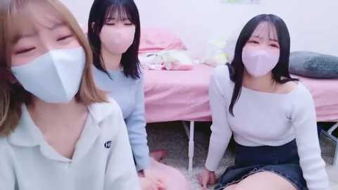 Video of three young Asian women in pink surgical masks, seated on a carpeted floor in a pink bedroom, wearing casual clothing, looking towards the camera.