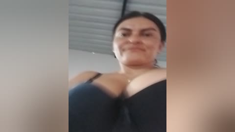 Media: Video of a smiling woman with medium skin tone, dark hair, and large breasts in a black bra, taken from a low angle. The background shows a white wall and a metal ceiling.