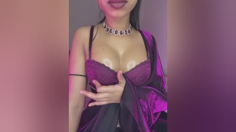 Media: Video of a woman with medium skin tone and long black hair, wearing a black satin robe, revealing a purple lace bra. She has a choker necklace with black and silver stones. Background is blurred with a soft pink and purple gradient.