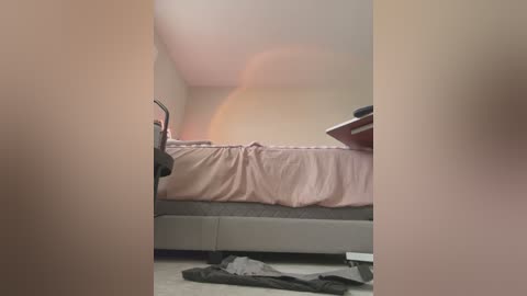 Media: A video of a minimalist bedroom with a bed covered in pink sheets, a black chair, and scattered clothes on the carpet. The room has beige walls and a small, uncluttered desk in the background.