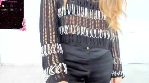 Media: Video of a woman with long blonde hair wearing a black, see-through knit top with white geometric patterns and high-waisted black shorts, standing indoors. Background includes a white wall and a pink flower arrangement.