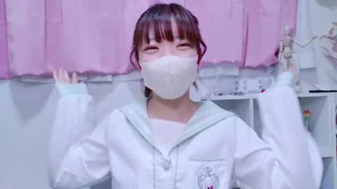 Media: Video of a young Asian woman with straight brown hair, wearing a white hoodie, white mask, and white gloves, smiling broadly, in a room with pink curtains and medical equipment.