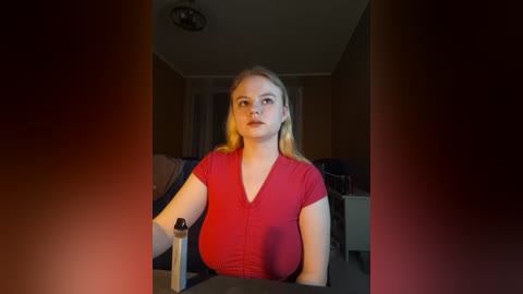 Media: Video of a young Caucasian woman with long blonde hair, seated in a dimly lit room, wearing a red V-neck top, gazing contemplatively, with a metallic object in her hand.