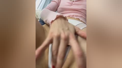 Media: A close-up video shows a woman's hand with red-painted nails reaching out, wearing a pink long-sleeved top, seated in a beige chair. The background is blurred, focusing on her hand and arm.