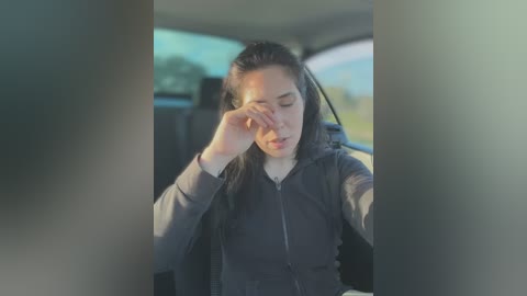 Media: A video captures an Asian woman with long black hair, wearing a dark jacket, shielding her eyes from the sun while sitting in a car. The background shows a blurred, sunny landscape.