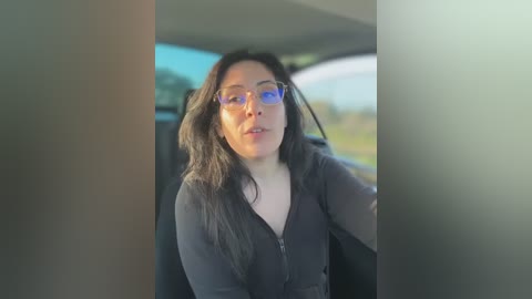 Media: Video of a middle-aged woman with long, dark hair and glasses, wearing a black jacket, sitting in a car, blurry background of a road and trees.