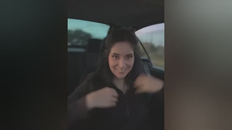 Media: A video of a smiling young woman in a car, with her hands gripping the steering wheel, wearing a black hoodie. The background features a blurred, dimly lit outdoor scene.