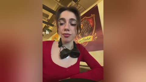 Media: Video of a young woman with pale skin, brown hair, wearing a red top and black headphones, sitting in a cozy room with wooden beams and a Hogwarts-themed poster in the background.