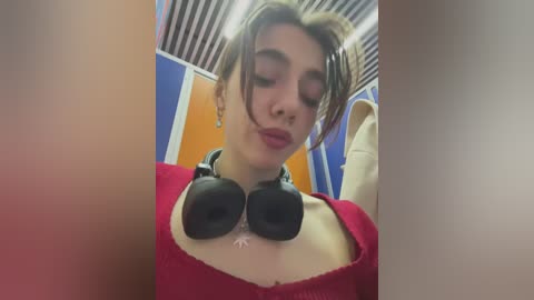 Media: Video of a young woman with light skin, short brown hair, wearing a red sweater, black headphones, and making a pouty face in a bathroom with blue and orange walls.