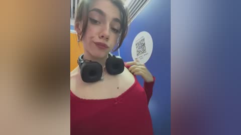 Media: Video of a young woman with short, light brown hair and fair skin, wearing a red top and black headphones. She holds a circular sign with text in a bathroom with blue and orange walls.