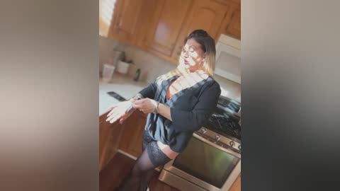 Media: Video of a curvy, light-skinned woman with long, blonde hair in a black blouse, revealing cleavage, black lace panties, and thigh-high stockings, standing in a kitchen with wooden cabinets, white countertops, and a stove.