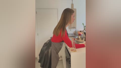 Media: A video of a Caucasian woman with straight, light brown hair, wearing a red top and gray apron, leaning over a white kitchen island, possibly cleaning. The background features a modern, minimalist kitchen with white walls and a wooden pendant light.