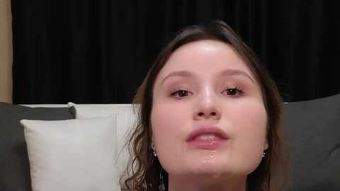 Media: A video of a young woman with light skin, shoulder-length brown hair, and a shiny face, possibly from sweat, wearing a white shirt, sitting on a grey couch with a white pillow, in front of a black curtain background.