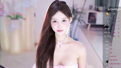 Media: Video of a young East Asian woman with long brown hair, wearing a strapless pink dress, a pearl necklace, and makeup, smiling softly, set in a modern indoor studio with blurred background.