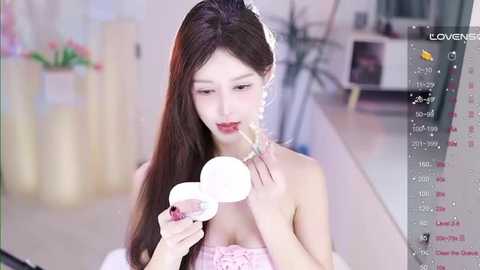 Media: A video of an East Asian woman with long, dark hair and fair skin, applying lipstick with a white brush. She wears a pink, strapless dress. The background shows a modern, minimalist room with potted plants and a glass table.