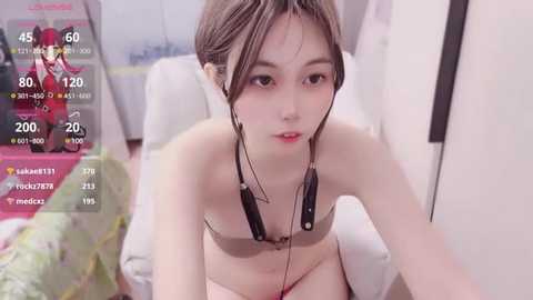 Media: A video of a young Asian woman with short, light brown hair, wearing a strapless bra, sitting on a bed. The room has a soft pink carpet and white walls.