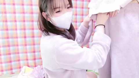 Media: A video of an Asian woman with long, straight black hair wearing a white face mask and a white jacket, assisting a person in a white bathrobe, set against a pastel checkered background.