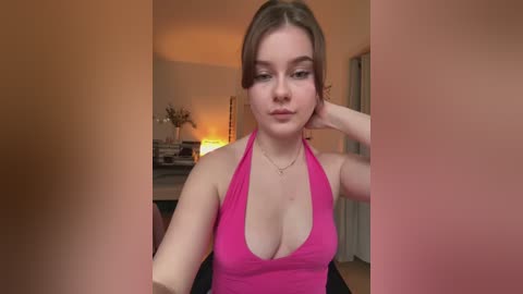 Media: Video of a young Caucasian woman with light skin, brown hair, and a slim build, wearing a bright pink halter top that accentuates her medium-sized breasts, in a warmly lit living room with dimly visible furniture.