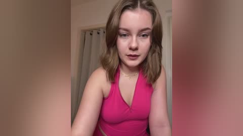 Media: Video of a young Caucasian woman with shoulder-length brown hair, wearing a bright pink halter top, standing indoors against a beige wall. She appears contemplative with a neutral expression.