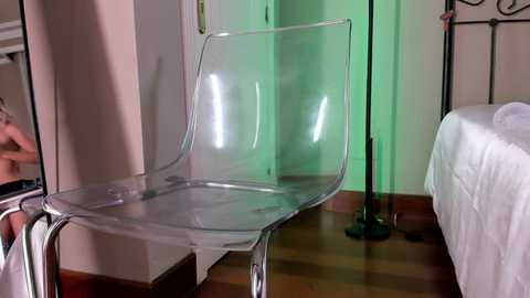 Media: Video of a modern, transparent acrylic chair in a minimalist bedroom with white walls, wooden floor, and a green-lit corner, featuring a bed with white sheets and a black metal frame.