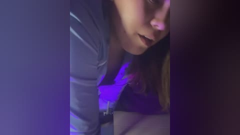 Media: Video of a young woman with light skin, light brown hair, and a light blue shirt, looking down, bathed in purple light.